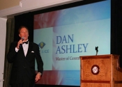 Dan Ashley speaks with microphone next to podium on stage with monitor display behind him at Diablo Country Club fundraiser.