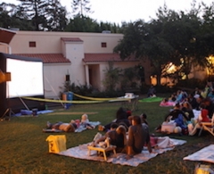 Crowds on picnic blankets watch outdoor movie on inflatable screen in large backyard.