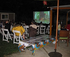Family and friends watch outdoor movie on inflatable screen at night in backyard.